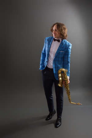 Full-length studio portrait of a young musician guy in a bright blue plaid jacket holding a saxophone.の写真素材