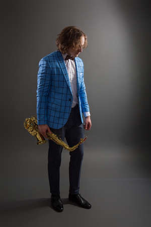 Full-length studio portrait of a young musician guy in a bright blue plaid jacket holding a saxophone.の写真素材