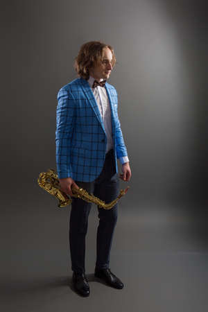Full-length studio portrait of a young musician guy in a bright blue plaid jacket holding a saxophone.の写真素材