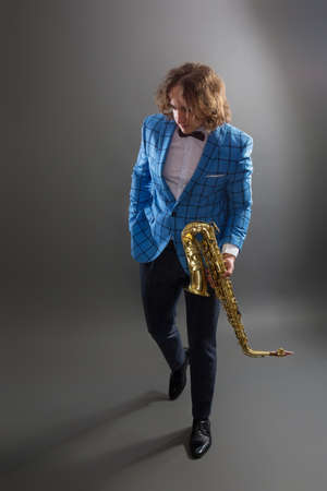 Full-length studio portrait of a young musician guy in a bright blue plaid jacket holding a saxophone.の写真素材