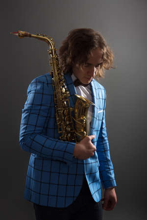 Half-length studio portrait of a young musician guy in a bright blue plaid jacket holding a saxophone.の写真素材