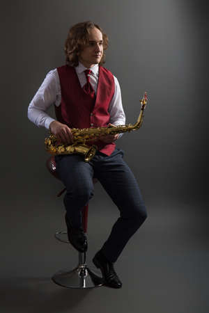 Young musician in a red vest sits and holds a saxophoneの写真素材