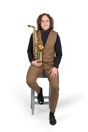 Young man in a black shirt and a light brown suit with a vest, sitting with her saxophone, isolated on white backgroundの写真素材