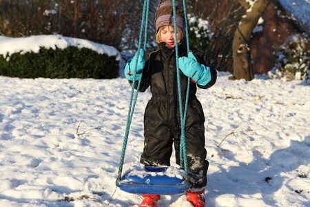 child by swing in snow. kid playing in garden on winter dayの写真素材