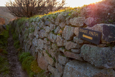 Wood sign with yellow letter to coast path hammered onto stone wall with view of path in the...