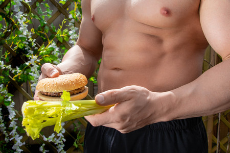 Fitness montage with healthy choice dilemma between fast food burger and healthy celery being held by man with chillselled physique, hard torso with pecs and abs.の写真素材