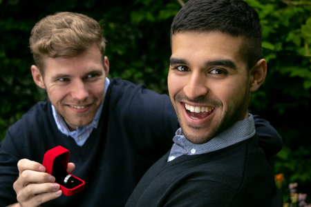 Engagement proposal betwen two gay men as one man proposes with an engagement ring in red box