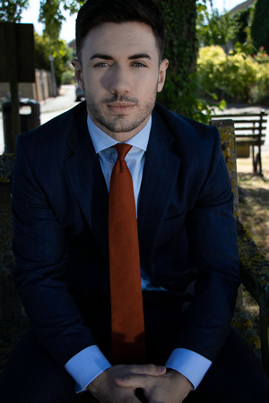 Handsome businessman with blue eyes and suit sits on village green bench at lunch timeの写真素材