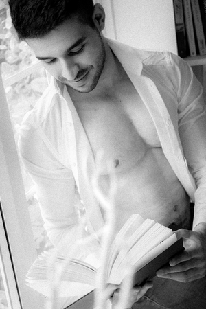 Man in sitting on window ledge with open shirt and pecs reading hardback bookの写真素材