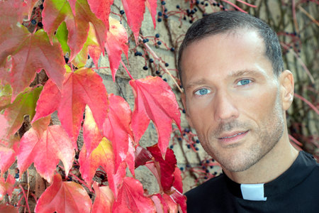 Good looking priest with visible collar poses for portrait next to red virginia creeper ivy