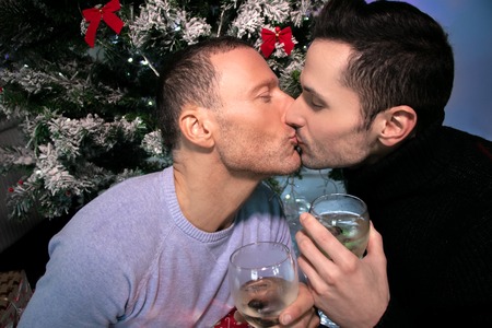 Happy male gay couple celebrating and kissing in front of Christmas tree with glasses of wineの写真素材