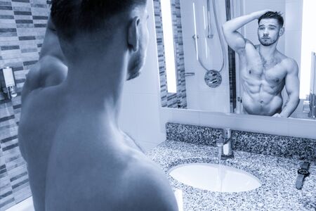 Attractive man with defined body looks at mirror reflection in bathroomの写真素材