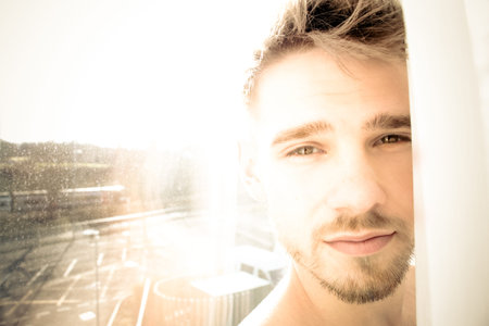 Portrait of handsome man lit by sunshine through window peeking out from behind curtainの写真素材