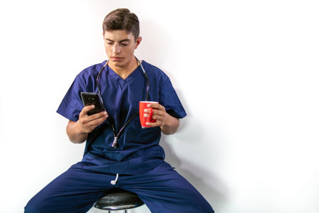 Handsome male doctor in blue scrubs, sitting on stool using phone while drinking from mug