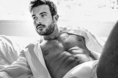 Handsome hairy muscular man with beard lying on bed in underwear and open shirt revealing sixpack abs and hairy chestの写真素材