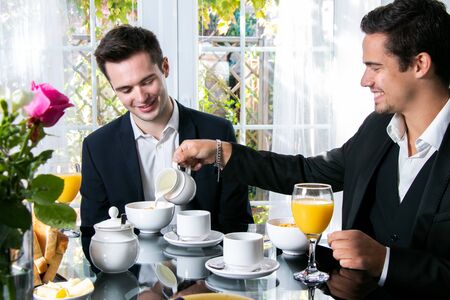 Friends or Gay couple sitting at table in front of patio doors eating breakfast and smilingの写真素材
