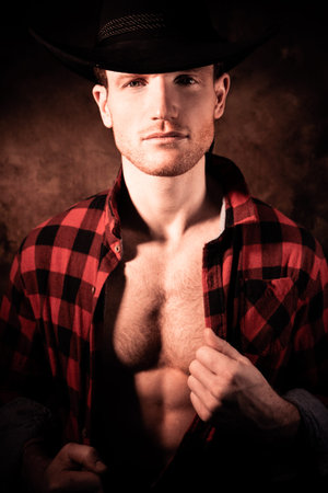 Portrait of handsome cowboy pulling chequered shirt open to reveal muscular pecs and defined sixpack...