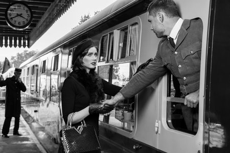 Vintage couple, man in uniform, woman in red dress, holding hands goodbye at train station as train...