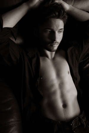 Portrait of attractive young man with beard, open shirt revealing chest, sitting in leather armchair, looking at cameraの写真素材