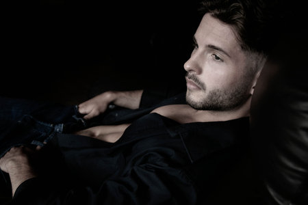 Portrait of attractive young man with beard, open shirt revealing chest, sitting in leather...