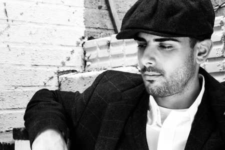 Handsome latino gangster dressed in shirt and jacket with flat cap sitting in front of white brick...