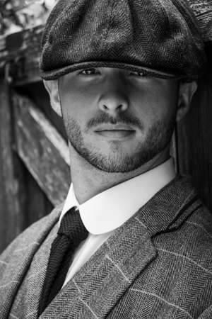 Handsome English gangster dressed in waistcoat and jacket sitting and looking at camera, leaning on gateの写真素材