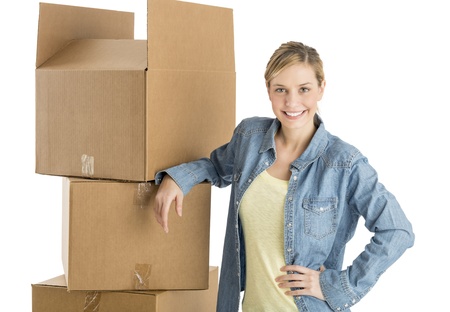 Portrait of happy young woman with hand on hip leaning on stacked cardboard boxes against white backgroundの写真素材