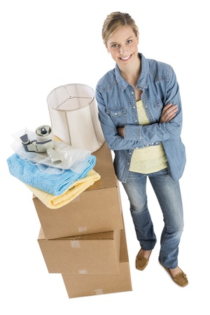 Full length portrait of confident young woman standing by stacked cardboard boxes with towels; adhesive tape and lamp shade on it over white backgroundの写真素材