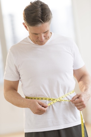 Front view of mature man measuring his waist while standing at health clubの写真素材
