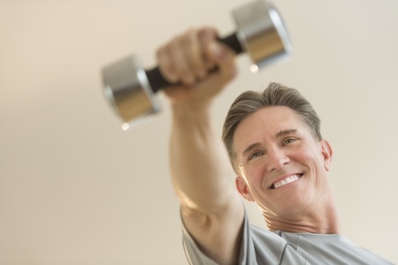 Low angle view of happy mature man lifting dumbbell against beige backgroundの写真素材