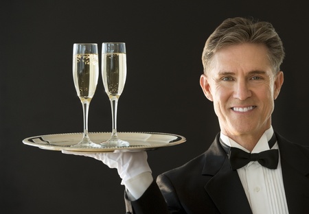 Portrait of happy mature waiter in tuxedo carrying serving tray with champagne flutes isolated on black backgroundの写真素材