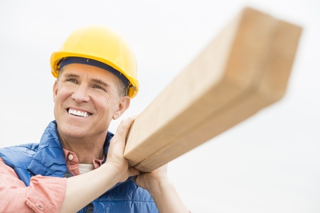 Happy construction worker looking away while carrying wooden plank against clear skyの写真素材
