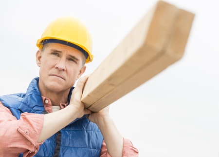 Thoughtful construction worker carrying wooden plank against clear skyの写真素材