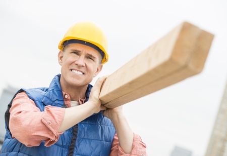 Male construction worker smiling while carrying wooden plank on shoulderの写真素材