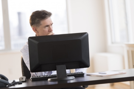 Thoughtful mature businessman looking away while sitting at computer desk in officeの写真素材