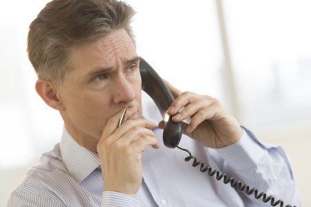 Thoughtful mature businessman looking away while using landline phone in officeの写真素材