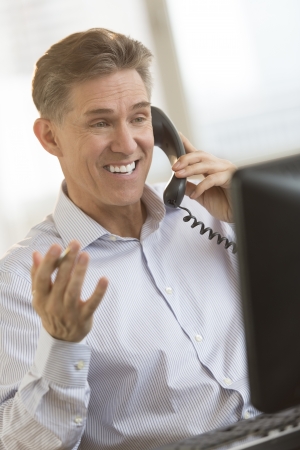 Happy mature businessman communicating on telephone while looking at computer monitor in officeの写真素材