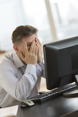 Frustrated mature businessman with hands on face looking at computer at desk in officeの写真素材