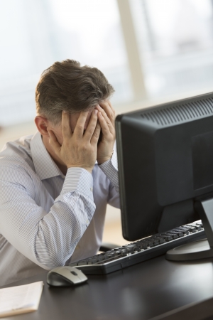 Tired mature businessman with hands on face leaning on computer desk in officeの写真素材