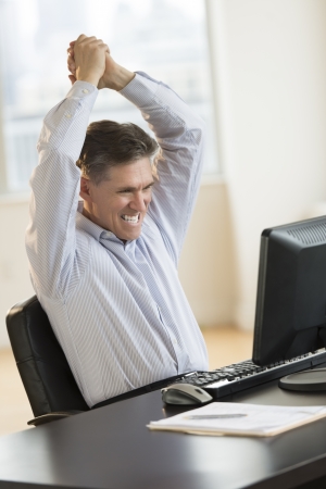Successful mature businessman with arms raised using computer at desk in officeの写真素材