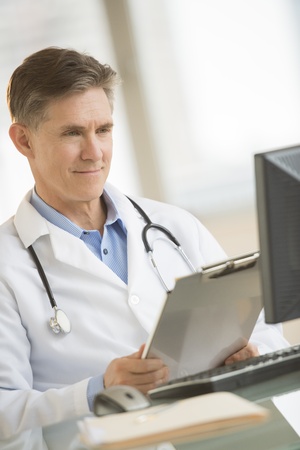 Mature male doctor smiling while looking at computer in clinicの写真素材