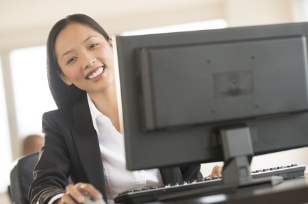Portrait of happy mid adult Asian businesswoman using computer at desk in officeの写真素材