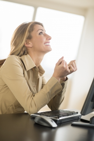 Happy young businesswoman looking up while sitting at desk with computer in officeの写真素材