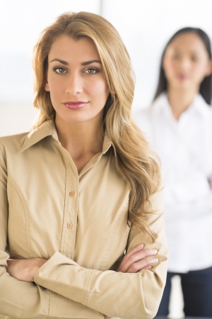 Portrait of confident businesswoman standing arms crossed at office with female colleague in backgroundの写真素材