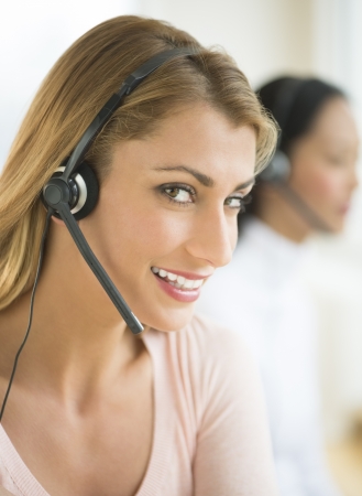 Close-up of portrait of happy female customer service representative wearing headset with colleague in backgroundの写真素材