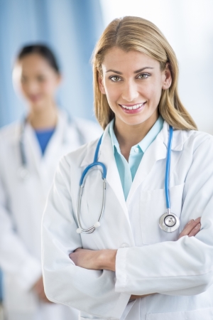 Portrait of happy young female doctor standing arms crossed at clinic with colleague in backgroundの写真素材