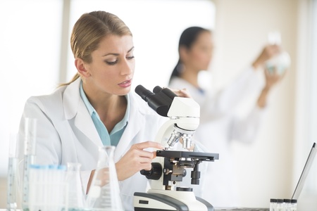 Young female scientist using microscope while colleague working in backgroundの写真素材