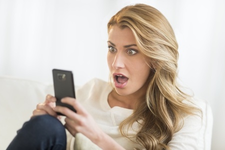 Shocked young woman looking at mobile phone while relaxing on sofa at homeの写真素材