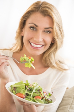 Portrait of happy young Caucasian woman eating vegetable salad while sitting on sofaの写真素材