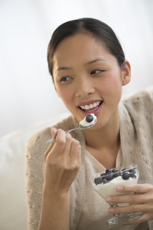 Beautiful young Asian woman looking away while eating yoghurt and olive at homeの写真素材